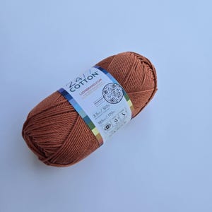 K&ouml;nnte beinhalten: Ein Kn&auml;uel 24/7 Cotton Garn in einem warmen, rotbraunen Farbton. Das Etikett zeigt das Lion Brand Logo, einen QR-Code und die Angaben: 100g und 170m. Das Garn ist fest gewickelt, ideal f&uuml;r Strick- oder H&auml;kelprojekte.