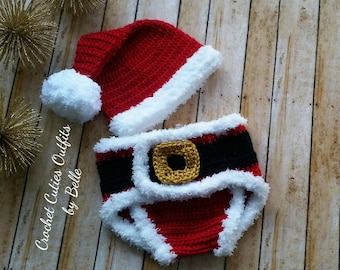 May include: A crocheted Santa Claus outfit for a baby, featuring a red and white hat with a white pom-pom and a red and white diaper with a black belt and gold buckle. "Crochet Cuties Outfits by Belle" is written on the left side of the image.