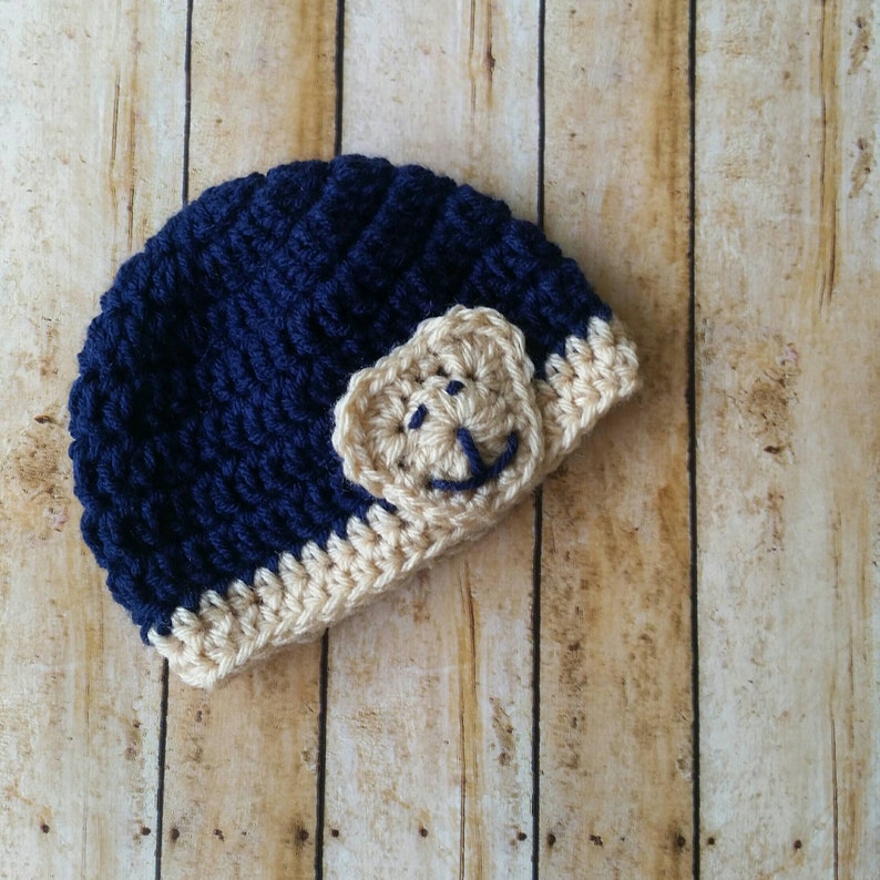 May include: A navy blue crocheted beanie with a tan crocheted bear face on the side. The bear has a smiling face.
