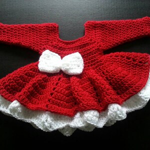 May include: A handmade red and white crochet baby dress. The dress features a red bodice and skirt with a white ruffled hem. Two white bows are centered on the bodice. The dress is approximately 12 inches long.