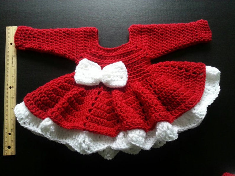 May include: A handmade red and white crochet baby dress. The dress features a red bodice and skirt with a white ruffled hem. Two white bows are centered on the bodice. The dress is approximately 12 inches long.