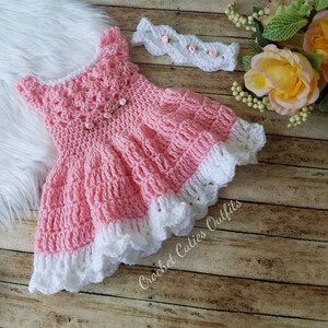 May include: A pink crocheted baby dress with a white lace trim and a matching white crocheted headband with pink flowers. The dress and headband are on a wooden surface with a white furry surface in the background.