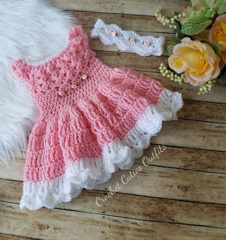 May include: A pink crocheted baby dress with a white lace trim and a matching white crocheted headband with pink flowers. The dress and headband are on a wooden surface with a white furry surface in the background.