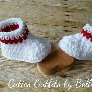 May include: A pair of white crocheted baby booties with red trim. The booties are sitting on a wooden heart-shaped surface.