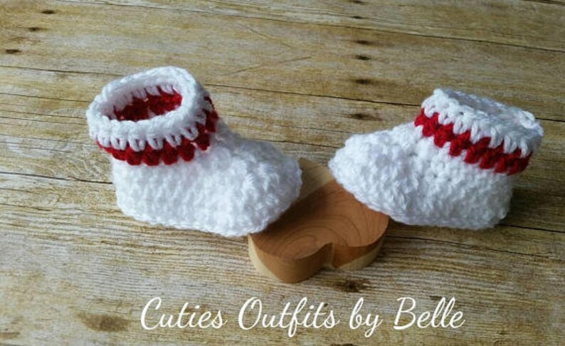 May include: A pair of white crocheted baby booties with red trim. The booties are sitting on a wooden heart-shaped surface.