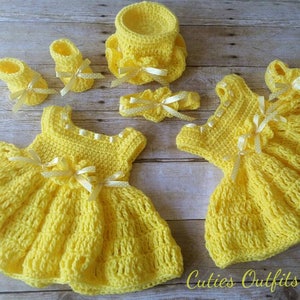 Peut inclure: Ensemble de v&ecirc;tements de b&eacute;b&eacute; au crochet jaune, comprenant deux robes, deux paires de chaussures, un bandeau et un petit panier. Chaque article est orn&eacute; de n&oelig;uds en ruban blanc. Le texte "Cuties Outfits by Belle" est visible.