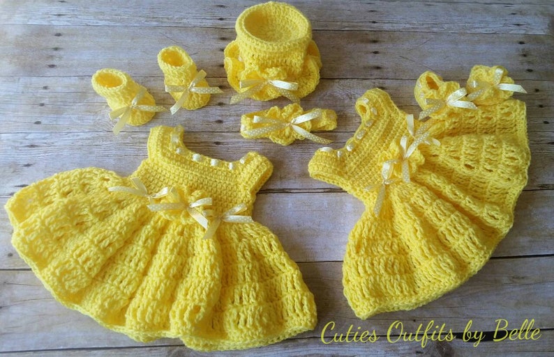 Peut inclure: Ensemble de v&ecirc;tements de b&eacute;b&eacute; au crochet jaune, comprenant deux robes, deux paires de chaussures, un bandeau et un petit panier. Chaque article est orn&eacute; de n&oelig;uds en ruban blanc. Le texte "Cuties Outfits by Belle" est visible.