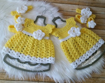 Könnte beinhalten: Zwei gelbe Häkelkleider für Babys mit weißem Besatz und Blumen. Die Kleider hängen an Kleiderbügeln und haben den Text "Crochet Cuties Outfits by Belle" unten.