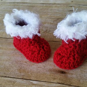 May include: A pair of red crocheted baby booties with white fur trim. The booties are on a wooden surface.