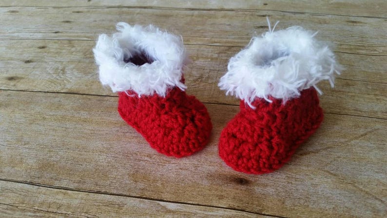 May include: A pair of red crocheted baby booties with white fur trim. The booties are on a wooden surface.