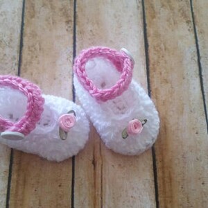 May include: A pair of white crocheted baby booties with pink trim and pink flower embellishments.