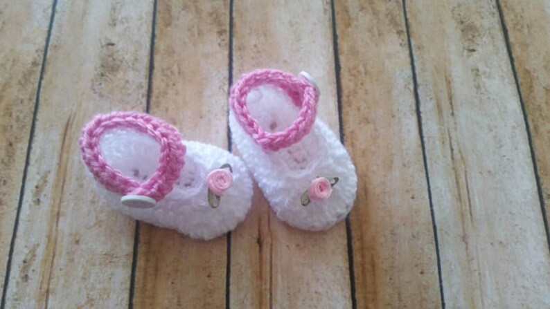 May include: A pair of white crocheted baby booties with pink trim and pink flower embellishments.