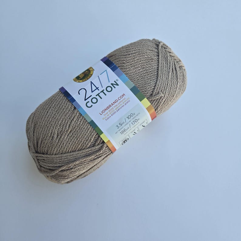 K&ouml;nnte beinhalten: Ein Kn&auml;uel Lion Brand 24/7 Cotton Garn in einem neutralen Beige. Das Etikett zeigt den Markennamen, die Garnst&auml;rke und die Faserzusammensetzung. Das Garn ist 100g und 170m. Ideal f&uuml;r Strick- und H&auml;kelprojekte.