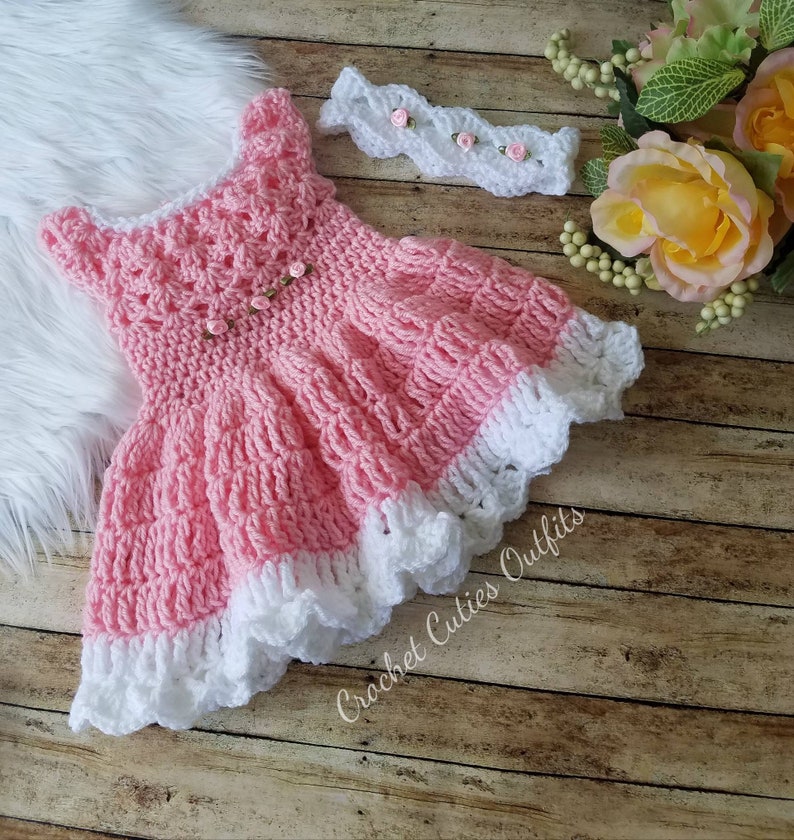 May include: A pink crocheted baby dress with a white lace trim and a matching white crocheted headband with pink flowers. The dress and headband are on a wooden surface with a white furry surface in the background.