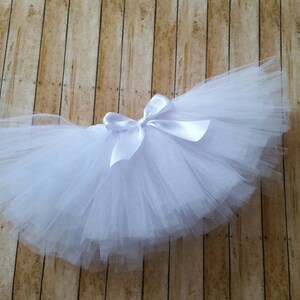 Peut inclure: Un tutu blanc avec un n&oelig;ud en satin. La jupe est faite de couches de tulle et est con&ccedil;ue pour les enfants. Le tutu est pr&eacute;sent&eacute; sur une surface en bois.