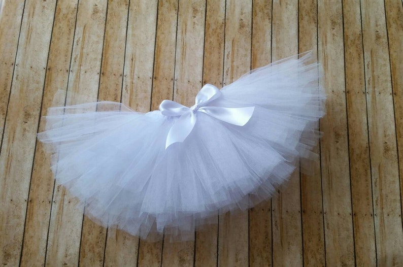 Peut inclure: Un tutu blanc avec un n&oelig;ud en satin. La jupe est faite de couches de tulle et est con&ccedil;ue pour les enfants. Le tutu est pr&eacute;sent&eacute; sur une surface en bois.