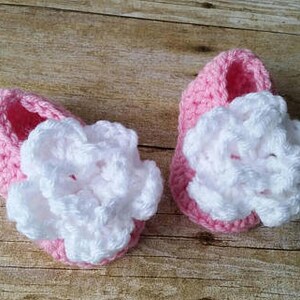 May include: A pair of pink crocheted baby shoes with white crocheted flowers on top.