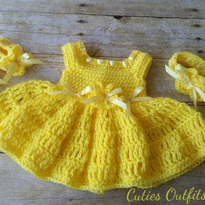 Peut inclure: Ensemble de v&ecirc;tements pour b&eacute;b&eacute; au crochet jaune fait &agrave; la main, comprenant une robe, des chaussures et un bandeau. La robe pr&eacute;sente une jupe &eacute;vas&eacute;e et est orn&eacute;e de petits n&oelig;uds. L'ensemble est pr&eacute;sent&eacute; sur une surface en bois. Le texte "Cuties Outfits by Belle" est visible.
