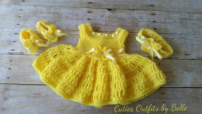 Peut inclure: Ensemble de v&ecirc;tements pour b&eacute;b&eacute; au crochet jaune fait &agrave; la main, comprenant une robe, des chaussures et un bandeau. La robe pr&eacute;sente une jupe &eacute;vas&eacute;e et est orn&eacute;e de petits n&oelig;uds. L'ensemble est pr&eacute;sent&eacute; sur une surface en bois. Le texte "Cuties Outfits by Belle" est visible.