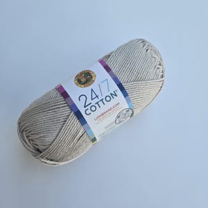K&ouml;nnte beinhalten: Ein Strang Lion Brand 24/7 Baumwollgarn in einem neutralen Beige. Das Garn ist fest gewickelt und hat ein Etikett mit dem Markennamen, der Garnart und der Website-Adresse. Das Etikett hat ein farbenfrohes Design mit den Worten "24/7 Cotton".