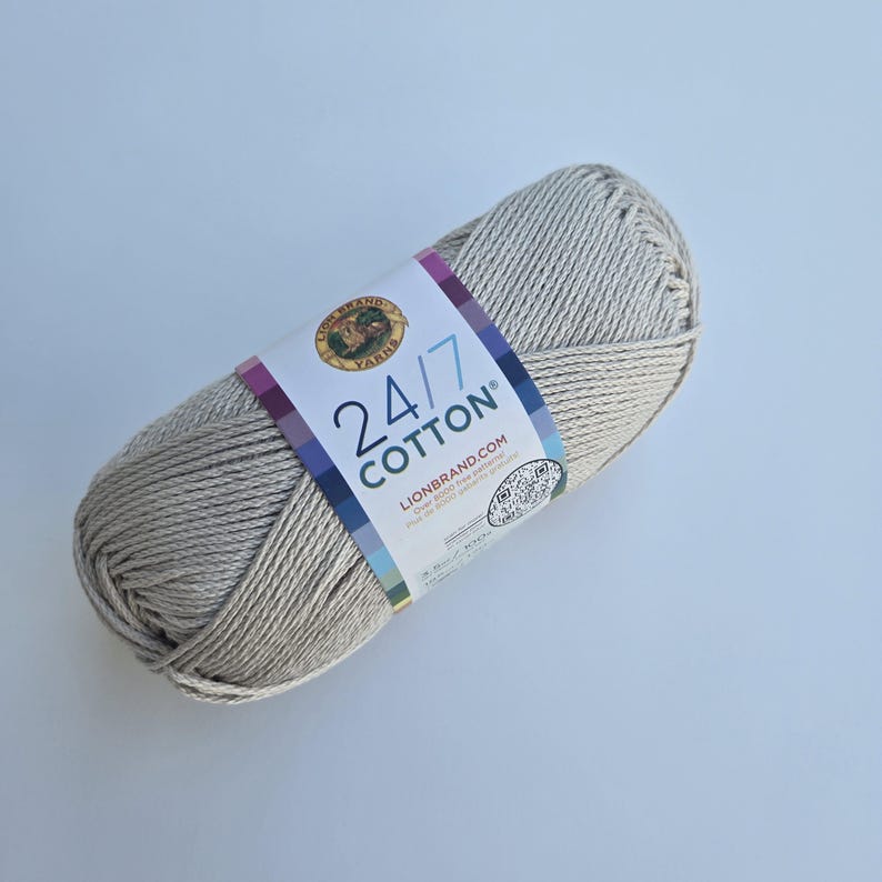 K&ouml;nnte beinhalten: Ein Strang Lion Brand 24/7 Baumwollgarn in einem neutralen Beige. Das Garn ist fest gewickelt und hat ein Etikett mit dem Markennamen, der Garnart und der Website-Adresse. Das Etikett hat ein farbenfrohes Design mit den Worten "24/7 Cotton".