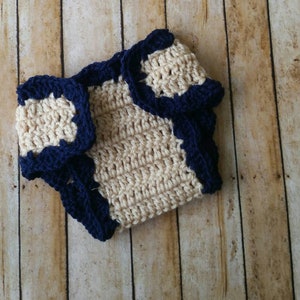 May include: A crocheted diaper cover in beige with a navy blue trim. The diaper cover is made with a simple stitch pattern and has a scalloped edge.