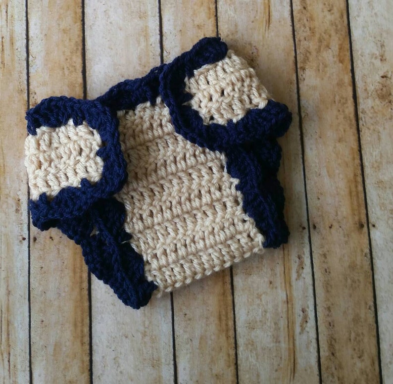 May include: A crocheted diaper cover in beige with a navy blue trim. The diaper cover is made with a simple stitch pattern and has a scalloped edge.