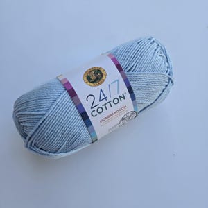 K&ouml;nnte beinhalten: Ein Kn&auml;uel hellblaues Lion Brand 24/7 Baumwollgarn. Das Garn ist mit einem Etikett versehen, das den Markennamen, die Garnart und die Website-Adresse enth&auml;lt. Das Garn hat eine weiche, einfarbige Farbe und eignet sich f&uuml;r Strick- und H&auml;kelprojekte.