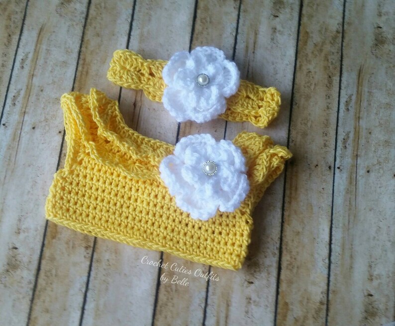 Peut inclure: Ensemble de crochet jaune pour b&eacute;b&eacute;, comprenant un haut et un bandeau. Le haut est orn&eacute; de volants, et les deux pi&egrave;ces sont d&eacute;cor&eacute;es de fleurs blanches avec des perles en leur centre. Le texte "Crochet Cuties Outfits by Belle" est visible.