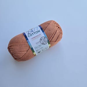 K&ouml;nnte beinhalten: Ein Kn&auml;uel pfirsichfarbener 24/7 Baumwollgarn. Das Garn ist mit einem Etikett mit dem Markennamen und Produktinformationen versehen. Das Etikett enth&auml;lt einen QR-Code und das Gewicht des Garns betr&auml;gt 100 g.