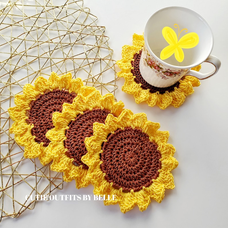 Pu&ograve; includere: Quattro sottobicchieri all'uncinetto a forma di girasole, di colore giallo e marrone. I sottobicchieri sono impilati uno sopra l'altro. I sottobicchieri sono su una superficie di metallo dorato.