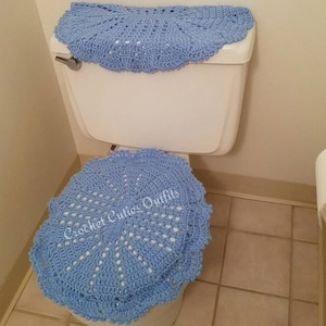 May include: A light blue crocheted toilet seat cover and a matching toilet tank cover. The covers are made with a delicate, lacy pattern. The text "Crochet Cuties Outfits" is visible on the seat cover.