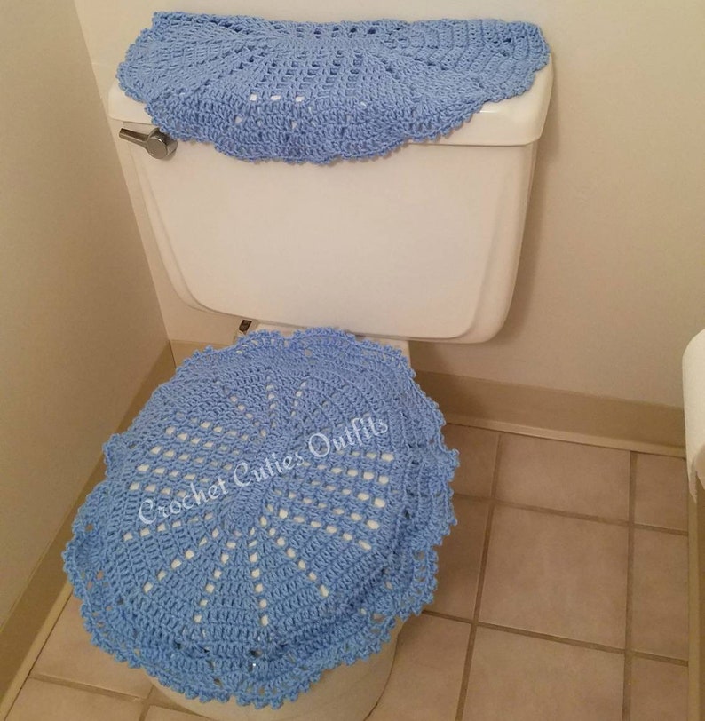 May include: A light blue crocheted toilet seat cover and a matching toilet tank cover. The covers are made with a delicate, lacy pattern. The text "Crochet Cuties Outfits" is visible on the seat cover.