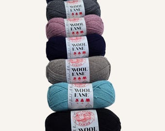Könnte beinhalten: Sieben Wollknäuel in verschiedenen Farben, darunter Marine, Grau, Mauve, Dunkelblau, Taupe, Türkis und Schwarz. Jedes Knäuel ist mit "WOOL EASE" und "Lion Brand Yarns" beschriftet. Die Wolle ist vertikal angeordnet und zeigt die Farbpalette.