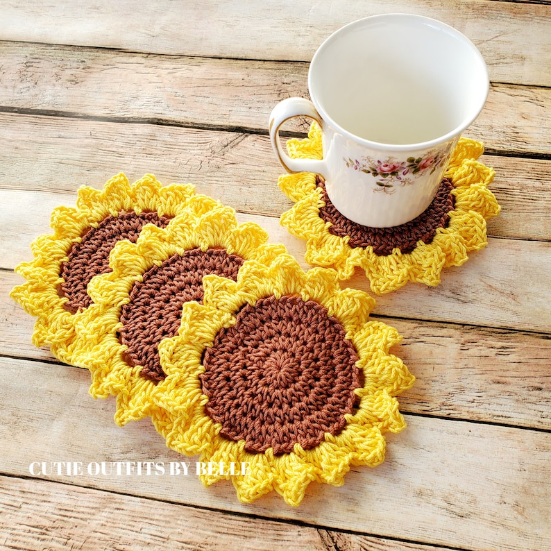 Pu&ograve; includere: Quattro sottobicchieri all'uncinetto a forma di girasole, di colore giallo e marrone. I sottobicchieri sono disposti in cerchio attorno a una tazza da t&egrave; bianca con motivi floreali.