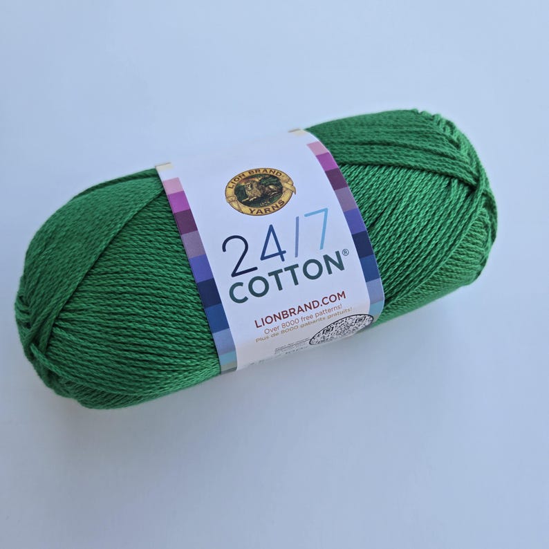 K&ouml;nnte beinhalten: Ein Strang gr&uuml;ne 24/7 Cotton Wolle. Die Wolle ist in einem leuchtenden Gr&uuml;nton gehalten und zu einer kompakten ovalen Form gewickelt. Ein Etikett umwickelt die Wolle und zeigt den Markennamen und Produktdetails.