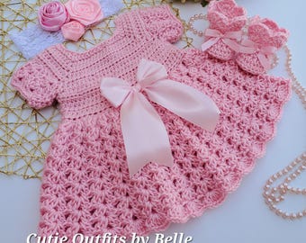 Può includere: Un vestitino da neonata in crochet rosa con un fiocco di raso rosa, una fascia abbinata con fiori rosa e un paio di scarpine in crochet rosa. "Cutie Outfits by Belle" è scritto in basso nell'immagine.