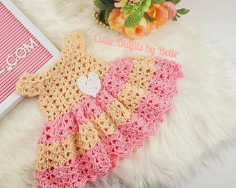 Puede incluir: Un vestido de bebé de ganchillo en color rosa y amarillo. El vestido tiene una aplicación de corazón blanco en la parte delantera y un dobladillo festoneado.