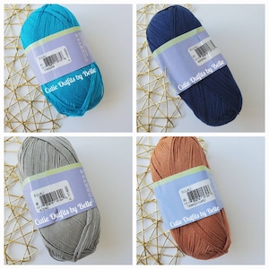May include: Four skeins of yarn in various colors: turquoise, navy blue, grey, and a light brown. Each skein has a label with the text "Cutie Outfits by Belle" and the yarn color name. The yarn is arranged on a gold wire mesh.