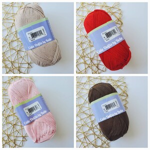 May include: Four skeins of yarn in various colors: beige, red, pink, and brown. Each skein has a label that reads "Cutie Outfits by Belle" and the yarn's color name. The yarn is displayed on a gold-colored, geometric patterned surface.