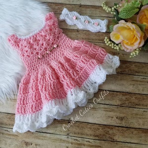 May include: A pink crocheted baby dress with a white lace trim and a matching white crocheted headband with pink flowers. The dress and headband are on a wooden surface with a white furry surface in the background.