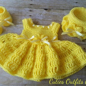 Peut inclure: Ensemble de v&ecirc;tements pour b&eacute;b&eacute; au crochet jaune vif fait &agrave; la main. L'ensemble comprend une robe, des chaussons et un bonnet, tous orn&eacute;s de d&eacute;licats n&oelig;uds en ruban. La robe pr&eacute;sente une jupe &eacute;vas&eacute;e et un corsage ajust&eacute;. Le texte "Cuties Outfits by Belle" est visible.