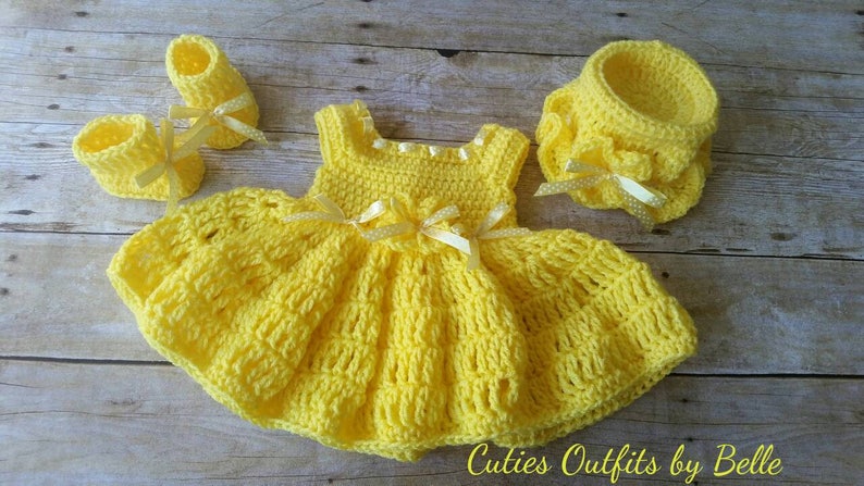 Peut inclure: Ensemble de v&ecirc;tements pour b&eacute;b&eacute; au crochet jaune vif fait &agrave; la main. L'ensemble comprend une robe, des chaussons et un bonnet, tous orn&eacute;s de d&eacute;licats n&oelig;uds en ruban. La robe pr&eacute;sente une jupe &eacute;vas&eacute;e et un corsage ajust&eacute;. Le texte "Cuties Outfits by Belle" est visible.
