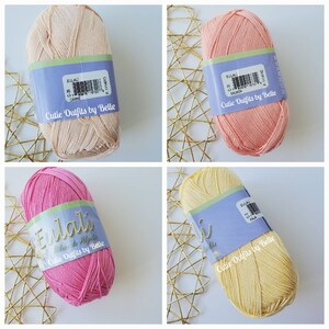 May include: Four skeins of yarn in various colors: light peach, salmon, hot pink, and pale yellow. Each skein has a label that reads "Eulali" and "Cutie Outfits by Belle". The yarn is displayed on a gold geometric patterned surface.