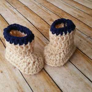 May include: A pair of crocheted baby booties in beige with a navy blue trim. The booties are made with a textured stitch and have a soft, cozy look.