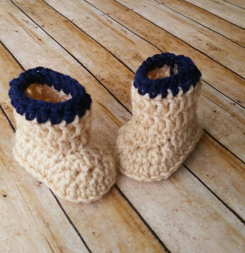 May include: A pair of crocheted baby booties in beige with a navy blue trim. The booties are made with a textured stitch and have a soft, cozy look.