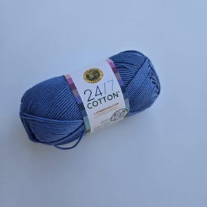 K&ouml;nnte beinhalten: Ein Kn&auml;uel blaue 24/7 Baumwollgarn. Das Garn ist mit einem Etikett versehen, auf dem "24/7 Cotton" und "LIONBRAND.com" steht. Das Garn hat eine einfarbige, mittelblaue Farbe. Der Hintergrund ist hellblau.