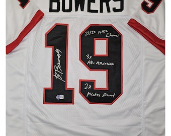 May include: White football jersey with the number 19 on the back. The name "BOWERS" is on the back of the jersey. The number 19 is signed with the name "B. Bowers" and includes the text "21/22 NATL Champ! 3x All-American 2x Mackey Award".