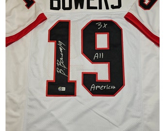 May include: White football jersey with black number 19, the name "BOWERS" on the back, and the text "3x All American" below the number. The jersey is signed "B. Bowers 19".