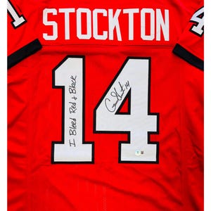 May include: Red jersey with the name "STOCKTON" in white block letters across the top. The number "14" is prominently displayed in white with a black outline. The jersey is signed and includes the text "I Bleed Red + Black".
